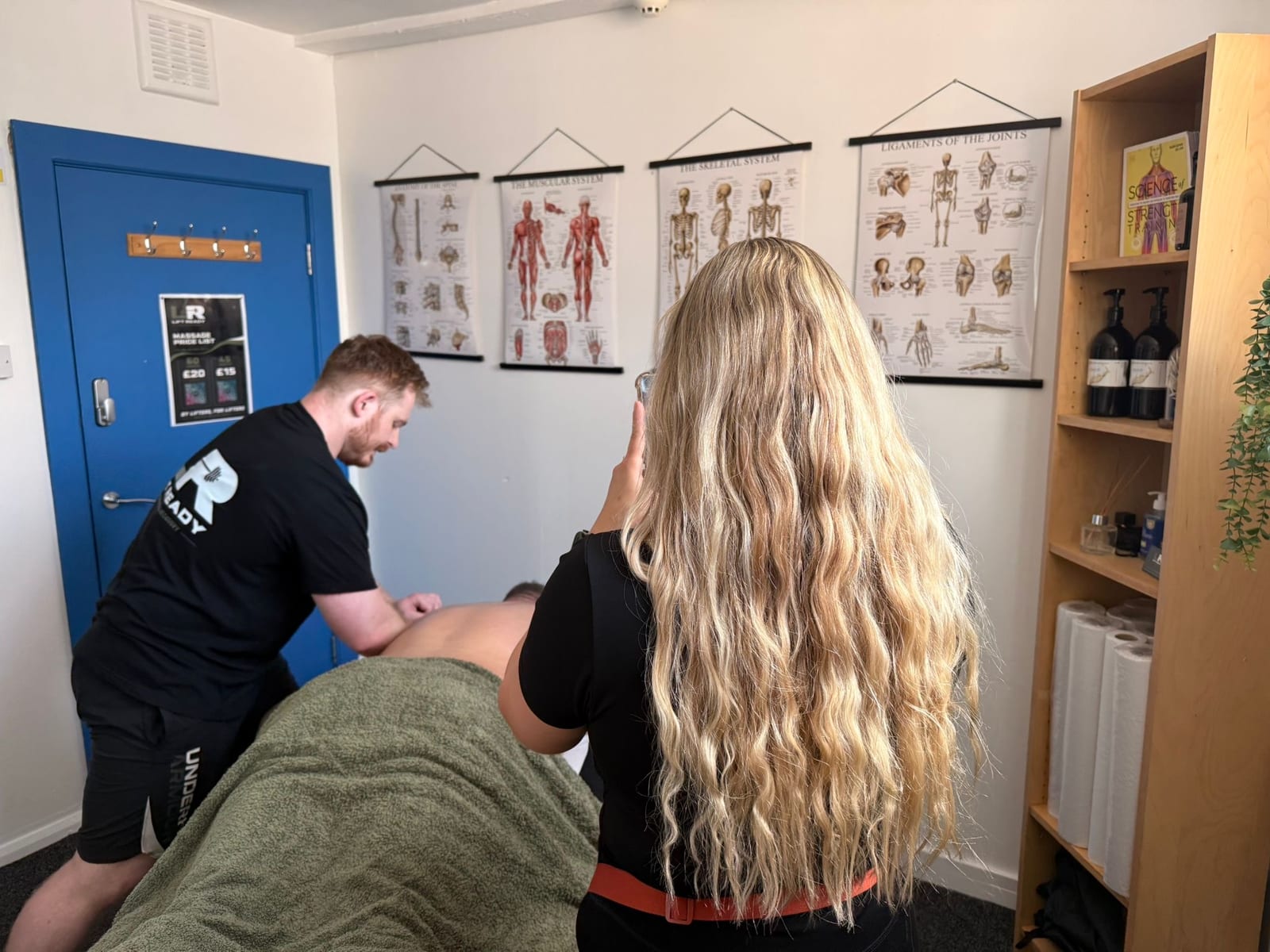Amelia from behind, phone raised, filming a therapist at work beneath a row of anatomy charts.